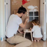 A man and child paint a cardboard house together on the floor, reflecting teamwork like planning a Nevada Living Trust. A man and child paint a cardboard house together on the floor, reflecting teamwork like planning a Nevada Living Trust.