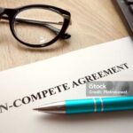 A document titled Non-Compete Agreement on a desk with black eyeglasses, a blue pen, and part of a calculator nearby. A document titled Non-Compete Agreement on a desk with black eyeglasses, a blue pen, and part of a calculator nearby.