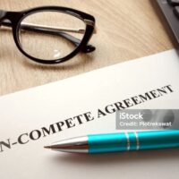 A document titled Non-Compete Agreement on a desk with black eyeglasses, a blue pen, and part of a calculator nearby. A document titled Non-Compete Agreement on a desk with black eyeglasses, a blue pen, and part of a calculator nearby.