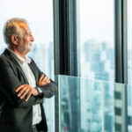 An older man in a suit stands, arms crossed, looking thoughtfully out large windows at a city skyline—reflecting on legacy. An older man in a suit stands, arms crossed, looking thoughtfully out large windows at a city skyline—reflecting on legacy.