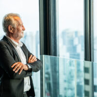 An older man in a suit stands, arms crossed, looking thoughtfully out large windows at a city skyline—reflecting on legacy. An older man in a suit stands, arms crossed, looking thoughtfully out large windows at a city skyline—reflecting on legacy.